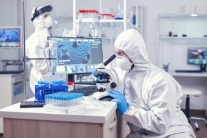 Zwei Menschen arbeiten in Schutzarbeitskleidung im Labor. Im Vordergrund sitzt eine Person vor einem Mikroskop, daneben stehen Reagenzgläser